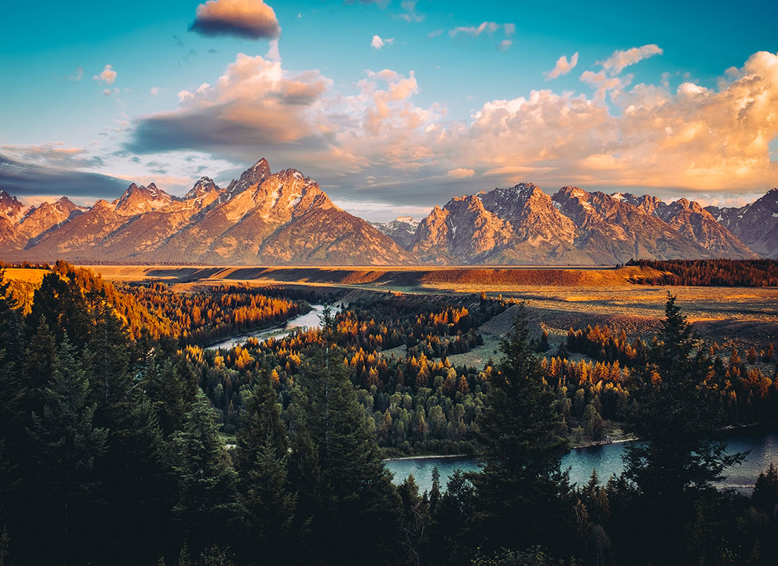 About Our Agency - Scenic Landscape of a River Surrounded by Green Trees with Views of the Mountains in the Background Against a Sunset Sky in Wyoming