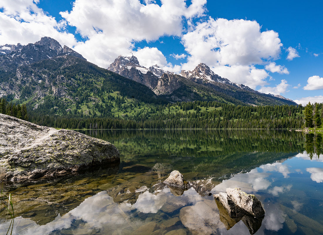 Contact - Scenic View of Rocks Along a Lake with Cloud and Tree Reflections in the Water in a State Park in Wyoming with Mountains in the Background Against a Cloudy Blue Sky